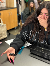 A high school student is covering her mouth in shock with one hand, while the other, open and holding a cockroach, is stretched out far away from her body.