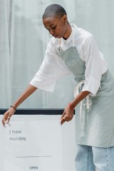 A woman in a white shirt, jeans, and gray apron is bending over and placing a sign that reads "I hate monday"